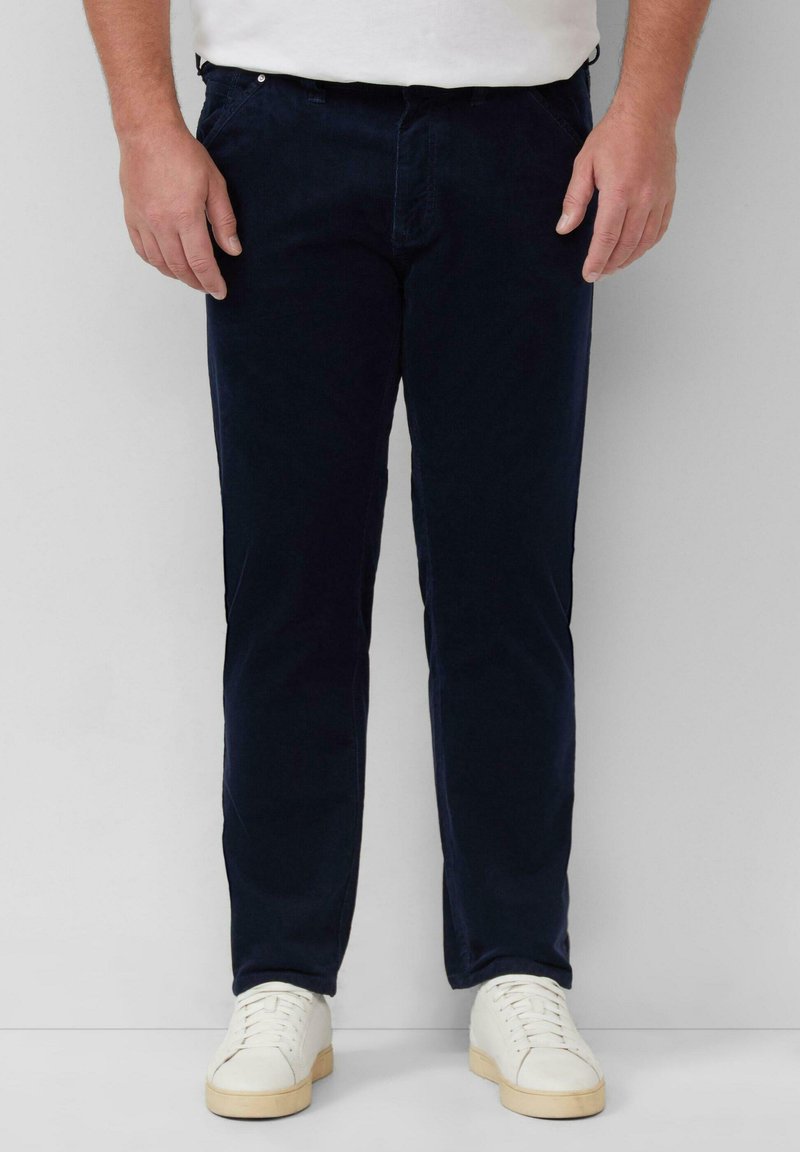 Man in een donkerblauwe broek en witte sneakers die tegen een lichtgrijze muur staat, met ontspannen handen langs de zijkanten.