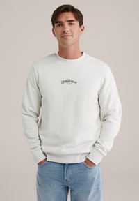 White sweatshirt with a round neckline and ribbed cuffs, featuring black embroidered text on the chest. Worn with light blue jeans.