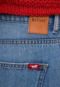 Modré džínsy z denimu s hnedou koženou etiketou s nápisom "MUSTANG" a červenou nášivkou s bielym logom koňa našitým na zadnej vrecku.