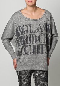 Szary sweter o luźnym kroju, z szerokim dekoltem i teksturowaną tkaniną. Zawiera wyraźne czarne napisy i subtelne przetarcia.