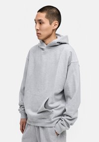 Grauer Kapuzenpullover aus weichem Material, lockere Passform, Kängurutasche, gerippte Bündchen und Saum sowie mit einer Kapuze mit Kordelzug. Model seitlich positioniert.