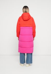 Cappotto imbottito con una parte superiore rossa e una parte inferiore rosa. Il design include un cappuccio e una vestibilità lunga e comoda. Pannelli in tessuto textured.