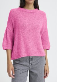 Femme portant un pull en tricot rose ample avec des manches arrivant aux coudes et un jean gris taille haute, sur un fond blanc uni.