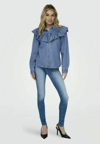 Denim srajca z velikimi volani na ramenih, modra barva, z gumbi spredaj, oblečena z svetlo modrimi ozkimi hlačami in črnimi visokimi petami.