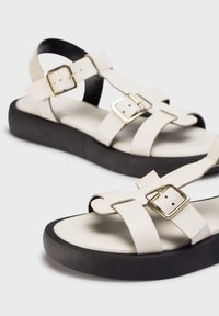 Sandalias de cuero blanco con suela de plataforma negra, con correas entrelazadas y hebillas doradas para el ajuste. Textura suave y diseño minimalista.