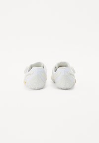 White athletic shoes with a textured mesh upper, cushioned heel, and rubber sole. Features a Velcro strap and slight gray accents.
