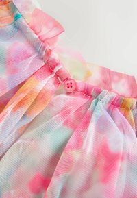 Gros plan sur un tissu transparent, coloré, tie-dye avec une taille froncée et un petit bouton rose au centre.