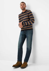 Uomo che indossa un maglione a righe marroni, blu e beige, jeans blu e stivali marroni con lacci, in piedi contro un muro bianco.