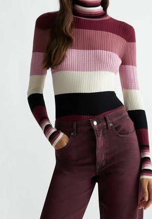 Femme portant un pull à col roulé côtelé avec des rayures horizontales en bordeaux, rose, blanc et noir, associé à un pantalon taille haute bordeaux.