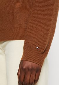 Bruine ribbeltrui met een gestructureerde afwerking en een klein logo-opdruk op de manchet, gecombineerd met crèmekleurige broeken.