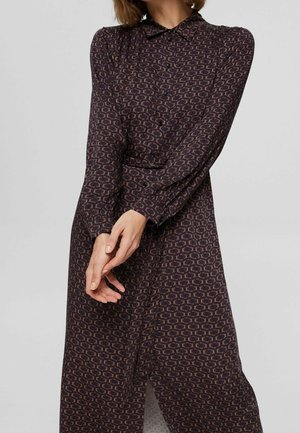 Frau trägt ein langärmliges, geknöpftes, dunkel gemustertes Kleid mit geometrischen Formen, steht mit sanft vor dem Körper gefalteten Händen.