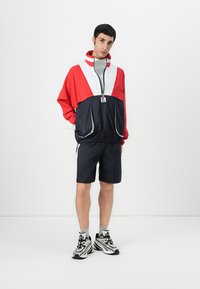 Rød, hvid og marinefarvet farveblokket vindjakke med høj krave, lynlås og to frontlommer, sammen med marinefarvede shorts og mønstrede sneakers.
