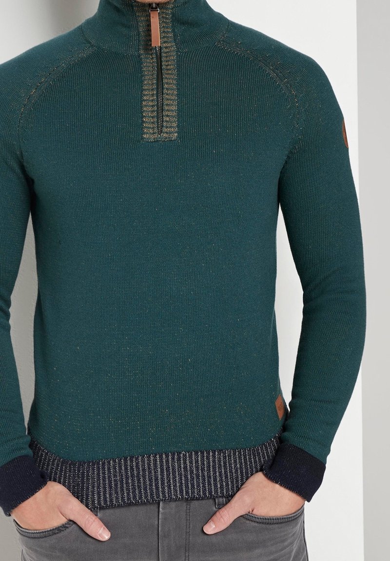 Maglione verde scuro a maglia con collo alto, mezza cerniera e dettagli a righe blu navy su polsini e orlo. Presenta una toppa logo color sabbia sul lato.