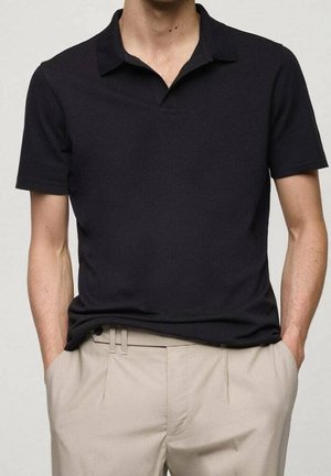 Mann trägt ein schwarzes kurzärmeliges Polohemd, in beige Hosen gesteckt, mit den Händen in den Taschen, steht vor einem schlichten Hintergrund.
