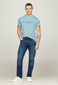 Camiseta de algodón azul claro con "TOMMY HILFIGER EST. 1985" impreso en negro, combinada con jeans de mezclilla oscura y zapatillas blancas.