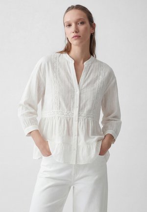 Kobieta z długimi włosami ma na sobie białą koszulę na guziki z koronkowymi detalami, rękawami do łokcia oraz białe spodnie, ręce w kieszeniach, neutralne tło.