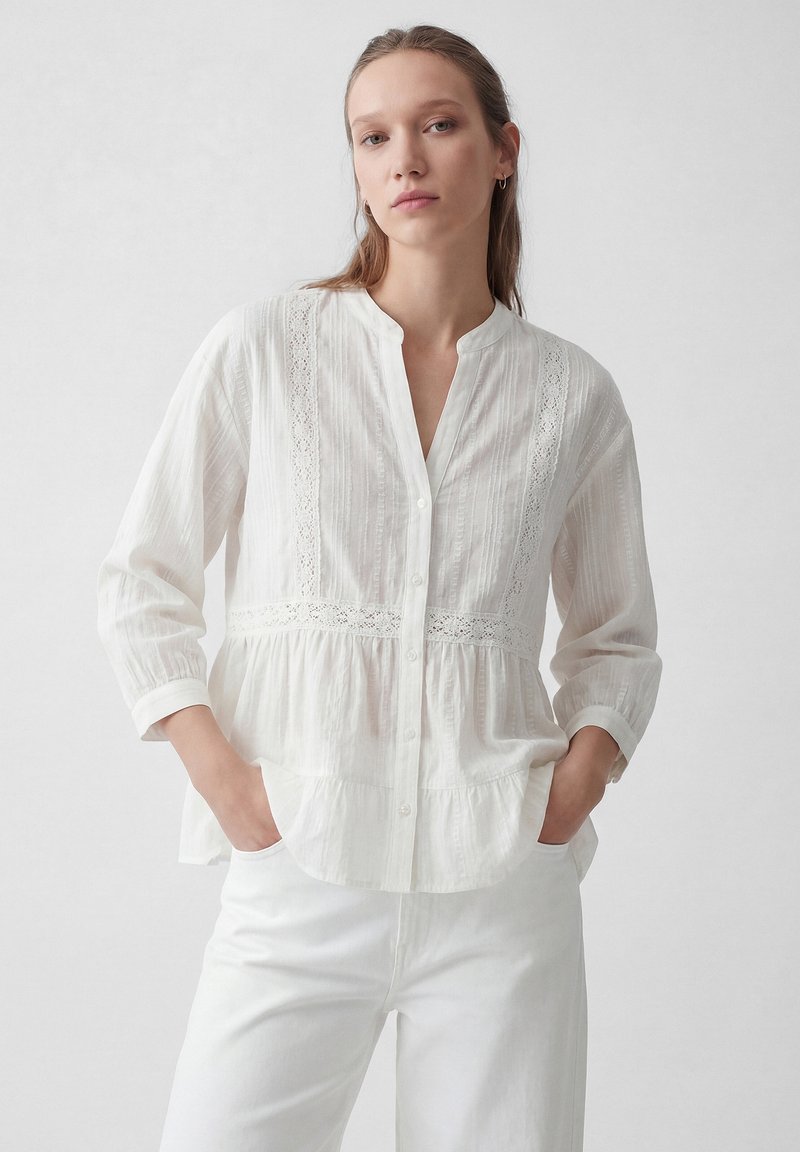 Kobieta z długimi włosami ma na sobie białą koszulę na guziki z koronkowymi detalami, rękawami do łokcia oraz białe spodnie, ręce w kieszeniach, neutralne tło.