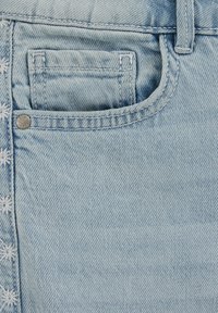 Tela vaquera de color azul claro con costuras visibles. Cuenta con un bolsillo delantero y un pequeño remache metálico en la apertura del bolsillo. Presenta bordado decorativo.