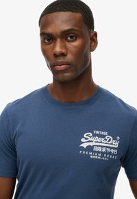 Mørkeblå bomulds t-shirt med et hvidt vintage SuperDry logo grafisk print foran, med rund hals og korte ærmer.