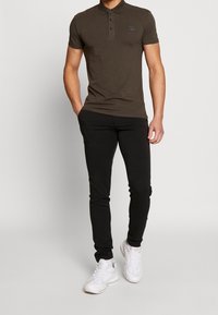 Tmavě olivově zelená polo tričko s úzkým střihem, s třemi knoflíky a malým logem na hrudi, v kombinaci s černými slim-fit kalhotami a bílými teniskami.