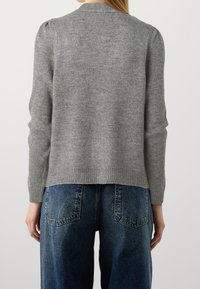 Osoba w szarej dzianinowej bluzie z bufkami i niebieskich dżinsach z wysokim stanem, widoczna z tyłu na jednolitym tle.