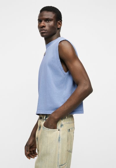 Νεαρός άνδρας με σκουρόχρωμο δέρμα, που φορά αμάνικο ανοιχτό μπλε μπλουζάκι και τζιν παντελόνι με ανοιχτόχρωμο ξέβαμμα, στέκεται με το ένα χέρι στην τσέπη, κοιτάζοντας μπροστά.