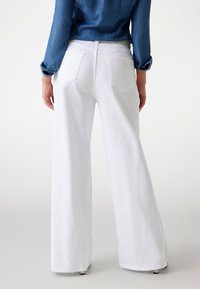 Osoba ubrana w białe, szerokie spodnie z wysokim stanem i długą niebieską koszulę dżinsową, stojąca tyłem do widza.