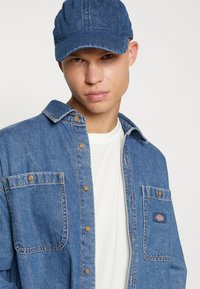 Τζιν πουκάμισο σε μεσαίο μπλε με γιακά, κουμπιά μπροστά και τσέπη αριστερά στο στήθος; φορεμένο με καπέλο σε ταιριαστό μπλε και λευκή μπλούζα από κάτω.