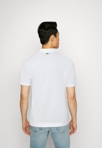 Polo bianco realizzato in cotone, con un colletto classico, maniche corte e un piccolo logo verde sulla parte posteriore del collo. Indossato con jeans blu.