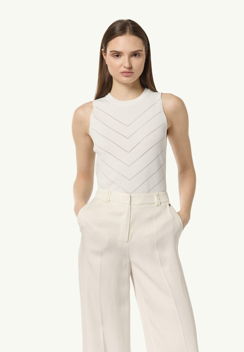 Weißer ärmelloser Body mit einem chevron Perforationsmuster, kombiniert mit hoch taillierten crèmefarbenen Hosen, die einen lockeren, geraden Schnitt zeigen.