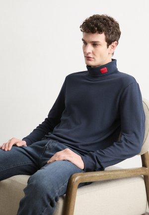 Jersey de cuello alto azul marino oscuro hecho de una tela suave, con mangas largas y una etiqueta roja en el borde del cuello. Combinado con unos vaqueros azules mientras está sentado.