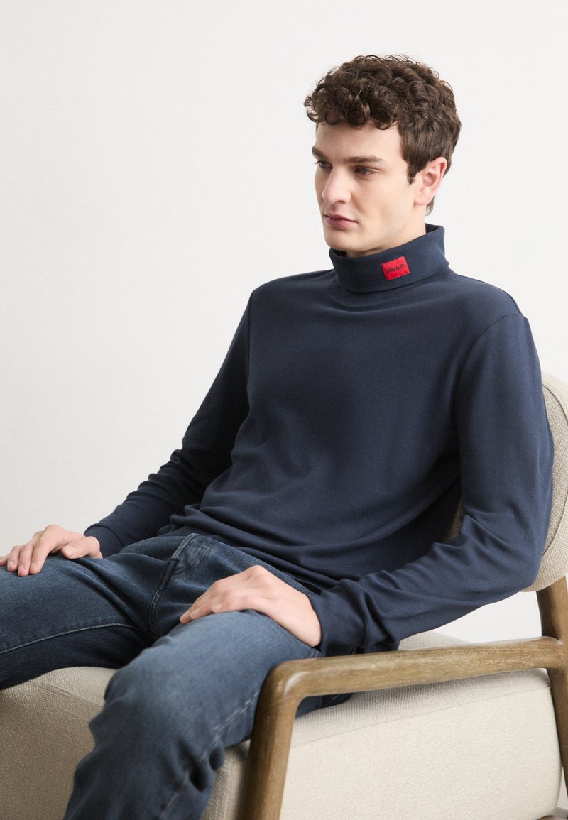 Donkerblauwe coltrui van gladde stof, met lange mouwen en een rood label aan de hals. Gekleed met blauwe jeans terwijl hij zit.