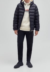 Hombre vistiendo una chaqueta acolchada negra sobre una sudadera beige con capucha, pantalones cargo negros con bolsillos laterales y botas negras con cordones frente a un fondo sencillo.