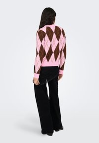 Rosa-brauner Argyle-Pullover mit langen Ärmeln, gerippten Bündchen und einer figurbetonten Passform, kombiniert mit schwarzen Hosen mit weitem Bein. Die Textur wirkt weich.