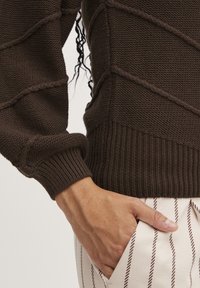 Brown textured knit sweater featuring a ribbed hem and sleeve cuffs, with raised line details. White and brown striped pants with pockets.