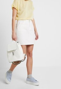 Bílý batoh s černými akcenty, nošený s žlutým topem a bílou sukní. Světle modré tenisky a šedé ponožky doplňují outfit.