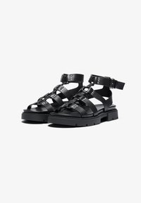 Sandali in pelle nera con larghe cinghie e borchie metalliche, dotati di una fibbia alla caviglia, suola spessa e design piatto per una maggiore stabilità.