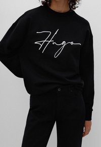 Schwarzes Sweatshirt mit lockerer Passform, mit einem weißen, geschwungenen "Hugo"-Logo. Kombiniert mit schwarzen Hosen. Weicher Stoff.