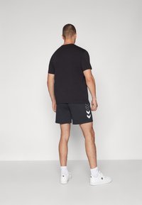 Svart kortärmad T-shirt i kombination med mörkgrå shorts med vita chevron-accenter, kompletterat med vita sneakers och ankelsockor.