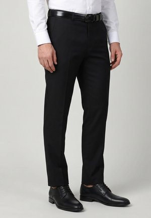 Mand iført sorte habitbukser, sort læderbælte, sorte pæne sko og en hvid langærmet skjorte foran en ensfarvet baggrund.