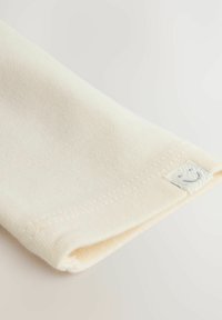 Kremkrāsas ribota auduma ar salocītu malu, kuram ir neliela izšūta birka ar gaiši pelēku smaidošu seju.