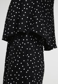 Robe noire avec des pois blancs, dotée d'un design supérieur superposé et de poches latérales. Fabriquée en tissu lisse.