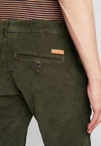 Ciemnozielone spodnie sztruksowe z tylną kieszenią zapinaną na guzik oraz beżową skórzaną metką marki przy talii.