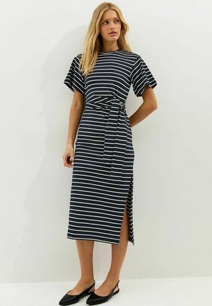 Vrouw die een midi-jurk met marineblauwe en witte strepen draagt, korte mouwen, zijsplit, tailleband en zwarte slingback platte schoenen, staand tegen een witte muur.