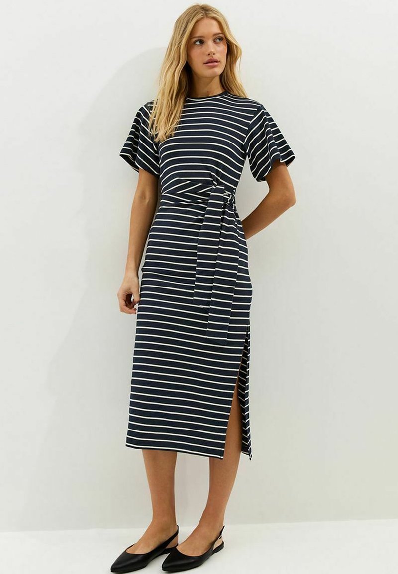 Mujer con vestido midi de rayas navy y blanco, de manga corta, con abertura lateral, cinturón en la cintura y zapatos planos negros con banda trasera, de pie contra una pared blanca.