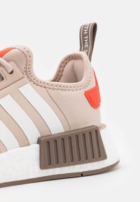 Beige athletic shoe with white stripes, orange accents, a textured brown heel cap, and a white cushioned sole. Features a pull tab.