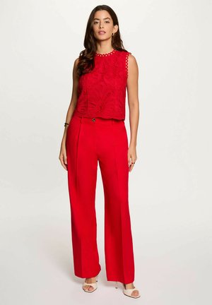 Donna in piedi che indossa un top rosso in pizzo senza maniche e pantaloni rossi a vita alta e gamba larga con tacchi bianchi aperti sulle dita, su sfondo chiaro.