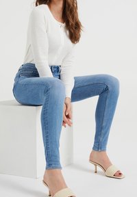 Světle modré skinny džíny v kombinaci s bílým pleteným kardiganem s texturou. Oblečení má volný střih a měkkou, lehkou látku.