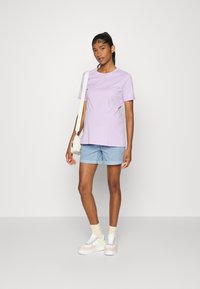 T-shirt de algodão lilás de mangas curtas, combinado com calções lisos azul claro. A modelo usa ténis em tons pastel e segura uma pequena bolsa.