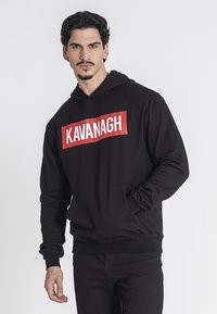 Čierna mikina s kapucňou vyrobená z mäkkého materiálu, má predné vrecko, rebrované manžety a veľké červené obdĺžnikové logo so slovom "KAVANAGH" v bielom texte.